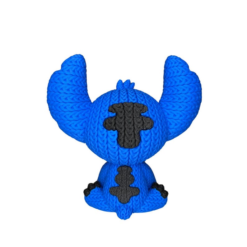 May include: A blue character figurine with large ears and black accents. The figurine has a textured, knitted appearance. The character is sitting with its arms and legs at its sides. The figurine is a popular character from a movie.