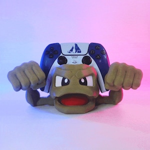 Può includere: Un originale supporto per controller per videogiochi a forma di personaggio dei cartoni animati marrone, con la bocca aperta e i pugni chiusi. Un controller blu e bianco con il logo di due lupi è posto sopra.