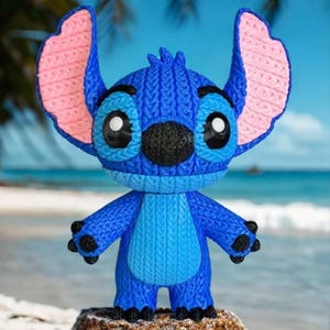 May include: A blue Stitch figurine with large ears, black nose, and big eyes. The figurine has a textured, knitted appearance with pink inner ears and black claws. It stands on a rock with a beach and ocean background.