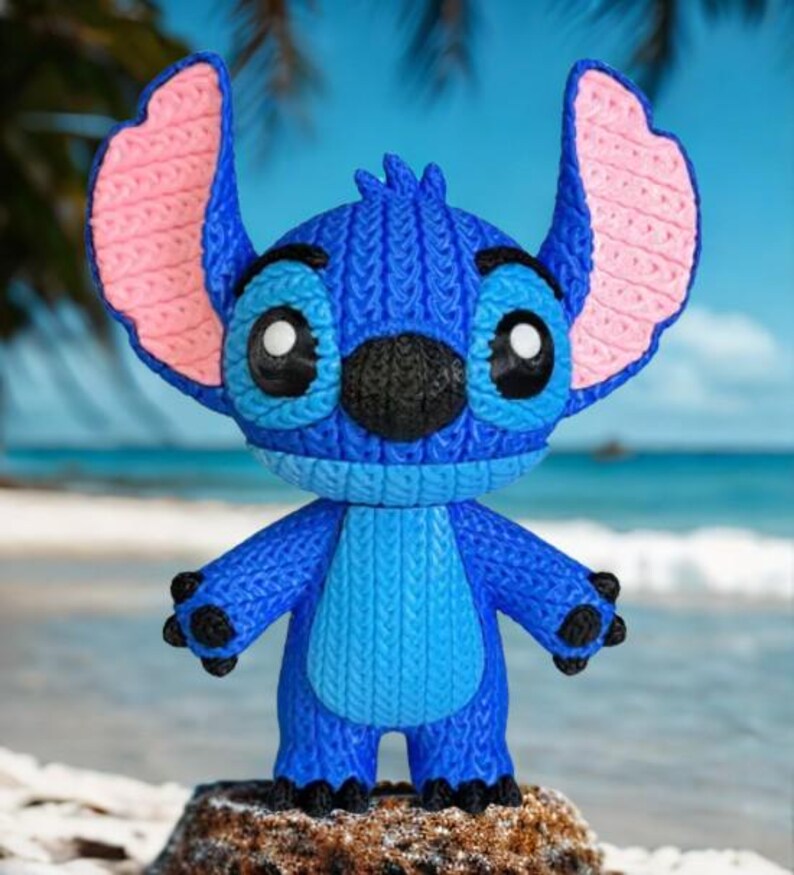 May include: A blue Stitch figurine with large ears, black nose, and big eyes. The figurine has a textured, knitted appearance with pink inner ears and black claws. It stands on a rock with a beach and ocean background.