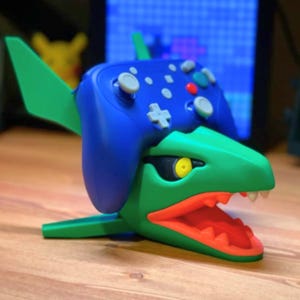 Puede incluir: Un soporte para mando verde y azul con forma de dragón con la boca abierta y un mando azul encima. El dragón tiene un ojo amarillo y dientes blancos. El mando tiene botones grises.