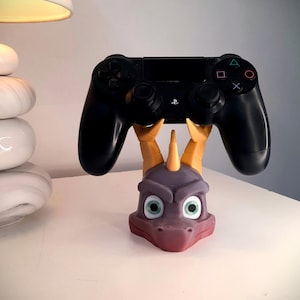 May include: A black PlayStation 4 controller rests on a purple and orange dragon-themed stand. The dragon has green eyes and a stern expression. The stand is designed to hold the controller securely.