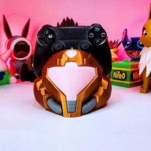 Puede incluir: Un soporte para mando de videojuegos marrón y dorado con forma de casco de personaje, con una visera rosa. Un mando negro descansa encima. Otras figuras de personajes están en el fondo.