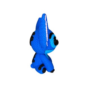 May include: A blue knitted character with black spots and large black eyes. The character has a prominent head with a pink stripe down the center. The figure is a handmade craft item.