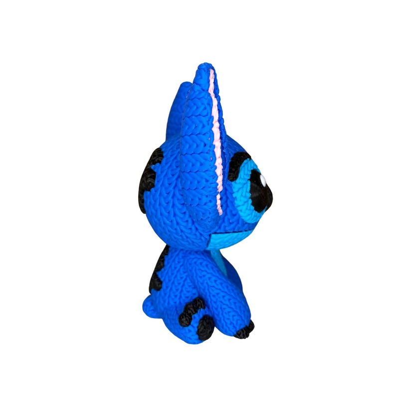 May include: A blue knitted character with black spots and large black eyes. The character has a prominent head with a pink stripe down the center. The figure is a handmade craft item.