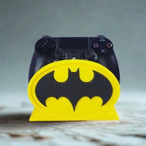 Puede incluir: Un mando negro de PlayStation 4 descansa en un soporte amarillo y negro con el tema de Batman. El soporte presenta el símbolo del murciélago. El mando es negro con botones y joysticks visibles. El soporte es amarillo con un símbolo de murciélago negro.