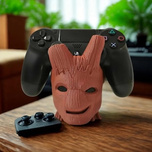 Puede incluir: Un soporte de mando marrón con forma de Groot con un mando negro de PlayStation 4. El soporte tiene una cara sonriente y sujeta el mando de forma segura. Un pequeño mando negro está sobre la superficie de madera.