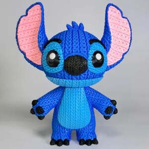 May include: A blue Stitch figurine with large ears, black nose, and big eyes. The ears have pink inner details. The figurine has a textured, knitted appearance. The figure is standing and has four black feet.