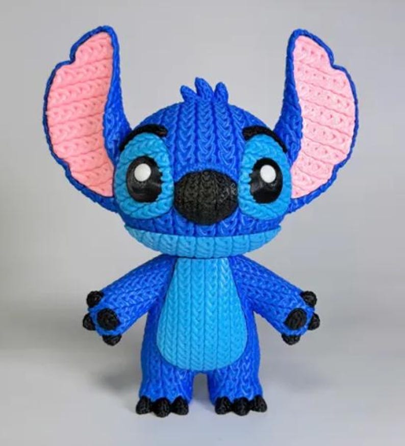 May include: A blue Stitch figurine with large ears, black nose, and big eyes. The ears have pink inner details. The figurine has a textured, knitted appearance. The figure is standing and has four black feet.