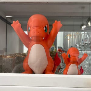 以下が含まれることがあります： 両手を広げた2体のオレンジ色のヒトカゲのフィギュア。大きなフィギュアは白いお腹と赤い尻尾をしています。小さなフィギュアは大きなフィギュアを模倣しています。どちらも緑色の目を持ち、ガラスのキャビネットの中に展示されています。