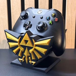 Può includere: Controller Xbox nero appoggiato su un supporto nero e oro stampato in 3D con un emblema stilizzato. Il supporto ha una base rettangolare e un design dettagliato. Il controller ha pulsanti colorati.