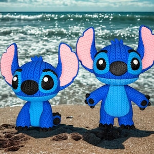 May include: Two blue cartoon character figurines with large ears and black eyes are on a sandy beach. The figurines have a knitted texture and pink inner ears. The ocean is visible in the background.