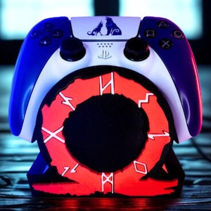 Puede incluir: Un mando de juegos azul y blanco con una base roja y negra. El mando tiene un diseño de lobo y un logotipo de PlayStation. La base presenta un diseño circular con detalles y símbolos rojos iluminados.