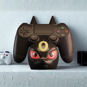 Puede incluir: Un soporte para mando negro con temática Umbreon con un mando de PlayStation 4 negro a juego. El soporte presenta la cara de Umbreon con ojos rojos y un anillo dorado. El mando descansa sobre el soporte.