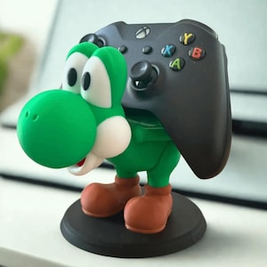 Può includere: Un supporto per controller a tema Yoshi verde che tiene un controller Xbox nero. Il supporto presenta una figura di Yoshi verde con dettagli bianchi, stivali marroni e una base nera. Il controller ha pulsanti colorati.
