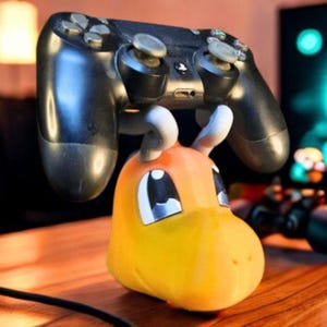 Puede incluir: Un mando negro de PlayStation 4 descansa sobre un soporte para mando con forma de dragón amarillo y naranja. El soporte tiene detalles blancos y un diseño de dibujos animados. El mando es negro y tiene un acabado brillante. El soporte está sobre una superficie de madera.