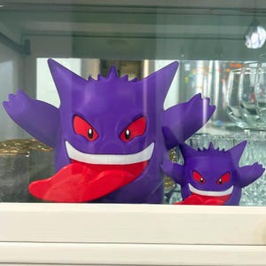 以下が含まれることがあります： 赤い目と舌を持つ2つの紫色のゲンガーのフィギュア。大きなフィギュアは腕を広げ、大きく笑っています。小さなフィギュアは大きなフィギュアを反映しています。フィギュアは白い棚に展示されています。