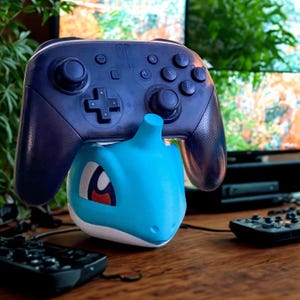 Puede incluir: Un soporte para mando con temática de Squirtle azul y blanco que sostiene un mando Switch Pro azul oscuro. El soporte está sobre una superficie de madera con otros mandos y un televisor al fondo.