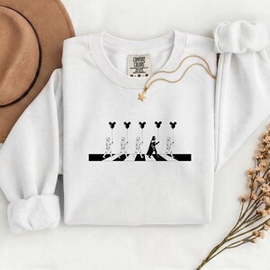 May include: White crewneck sweatshirt with a black and white graphic. The graphic depicts Stormtroopers and Darth Vader crossing a crosswalk, each holding a Mickey Mouse balloon. A gold necklace with a star pendant is also visible.