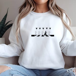 May include: White sweatshirt featuring a black and white graphic of characters crossing a crosswalk, each holding a Mickey Mouse-shaped balloon. The design is a parody of the iconic Abbey Road album cover.