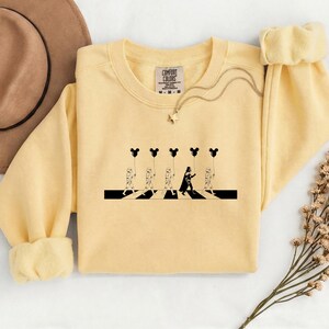 May include: A light yellow sweatshirt featuring a black and white graphic of characters crossing a street, holding Mickey Mouse-shaped balloons. The graphic is a parody of a famous album cover. The sweatshirt has a crew neck and long sleeves.