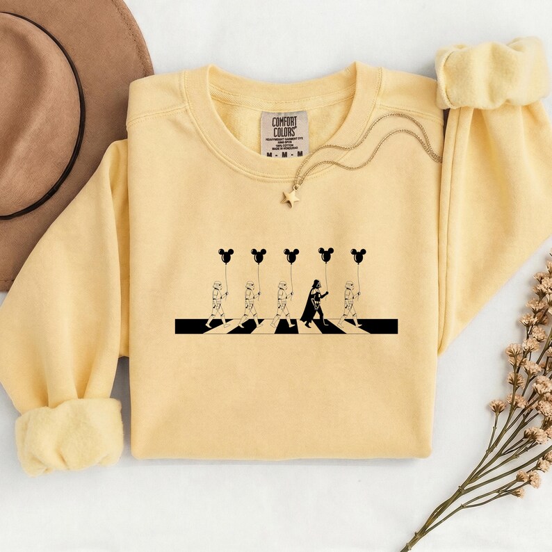 May include: A light yellow sweatshirt featuring a black and white graphic of characters crossing a street, holding Mickey Mouse-shaped balloons. The graphic is a parody of a famous album cover. The sweatshirt has a crew neck and long sleeves.