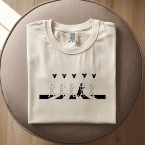May include: Cream-colored t-shirt featuring a black and white graphic. The design parodies the Beatles' Abbey Road album cover, with Star Wars characters crossing a crosswalk, holding Mickey Mouse balloons. The shirt has a Comfort Colors tag.