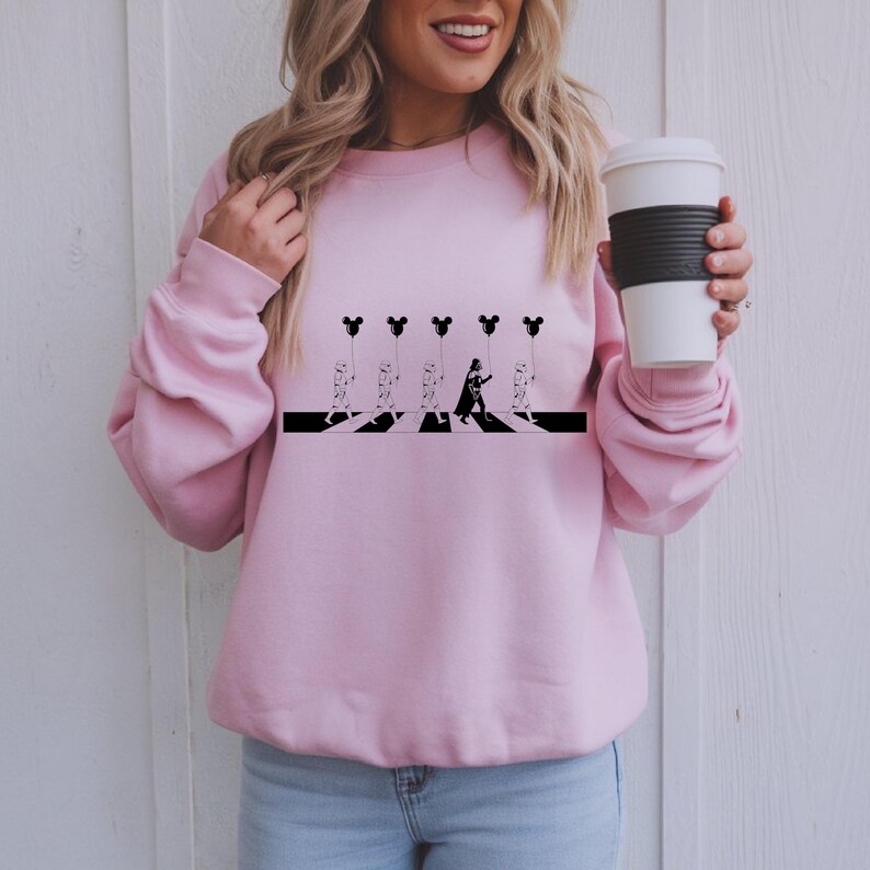 May include: A light pink sweatshirt featuring a black and white graphic. The graphic depicts a parody of the Abbey Road album cover, with characters and Mickey Mouse balloons. The sweatshirt is a crew neck style.
