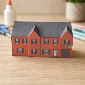 May include: A paper craft model of a two-story house with red walls, a dark gray roof, and dark gray shutters. The structure features multiple windows, a single front door, and a garage door on the right side, all rendered in a flat graphic style.