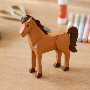 May include: A three-dimensional paper craft horse figure constructed from tan cardstock with a dark brown mane, tail, and hooves. The horse features geometric, angular facets and a printed eye detail, standing in a profile position on a flat surface.