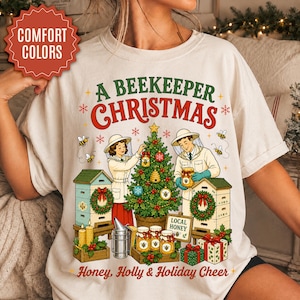 May include: An off-white t-shirt featuring a graphic with the text A BEEKEEPER CHRISTMAS, LOCAL HONEY, and Honey, Holly & Holiday Cheer. The design shows two beekeepers decorating a Christmas tree with honey-themed ornaments near beehives.