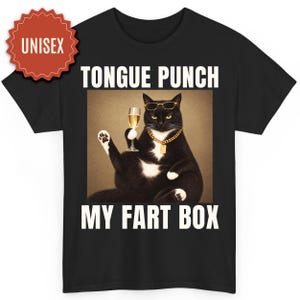 Könnte beinhalten: Ein schwarzes T-Shirt mit einer Grafik einer schwarz-weißen Katze, die eine Sonnenbrille und eine Goldkette trägt und ein Glas Champagner hält. Der Text TONGUE PUNCH MY FART BOX ist in Weiß über und unter der Katze gedruckt. Ein rotes UNISEX-Abzeichen befindet sich oben links.