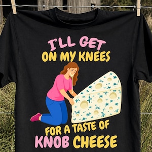 May include: A black t-shirt featuring a graphic of a person kneeling before a large wedge of blue cheese. The text reads I'LL GET ON MY KNEES FOR A TASTE OF KNOB CHEESE in pink and yellow lettering. The person wears a pink shirt and blue pants.