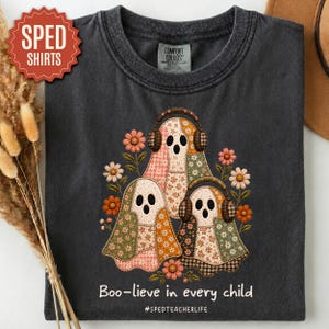May include: A charcoal gray crew neck t-shirt featuring a graphic of three ghosts wearing headphones, surrounded by floral patterns. The text reads "Boo-lieve in every child" and "#SPEDTEACHERLIFE" in white lettering below the ghost illustration.