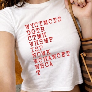 Puede incluir: Top corto blanco con texto rojo. El texto está dispuesto en formato vertical y escalonado. El top es de manga corta y parece estar hecho de un material suave y ligero. Una correa de bolso marrón es visible en el fondo.