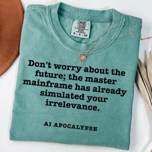 May include: A teal short-sleeved t-shirt with a crew neckline and a Comfort Colors label. The shirt features black text that reads: Don't worry about the future; the master mainframe has already simulated your irrelevance. AI APOCALYPSE.