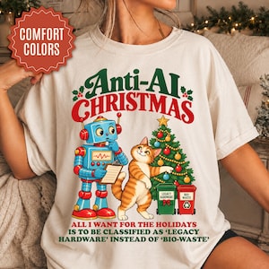 May include: A cream-colored t-shirt featuring a graphic of a blue robot and an orange tabby cat standing by a decorated Christmas tree. The text reads Anti-AI Christmas and All I want for the holidays is to be classified as Legacy Hardware instead of Bio-Waste.