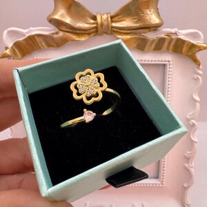 May include: A gold-colored ring with a four-leaf clover design, encrusted with small clear stones, and a heart-shaped pink stone. The ring is displayed in a teal gift box with a black interior. A gold bow is in the background.