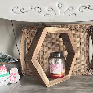 May include: A rustic, diamond-shaped wooden shelf holds a large candle jar with a black lid. The candle has a red wax base and a label with floral and text details. A woven tray and decorative snowmen are in the background.