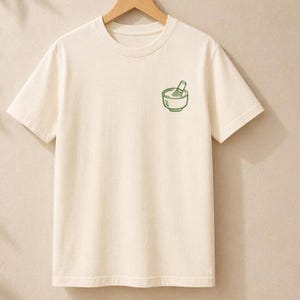 May include: A cream-colored short-sleeve t-shirt featuring a small, simple green line-art graphic on the left chest. The graphic depicts a bowl with a whisk inside, representing a matcha tea set. The shirt has a crew neckline and a straight hem.