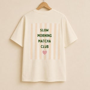 May include: A cream-colored short-sleeve t-shirt featuring a rectangular graphic on the back. The graphic displays vertical beige stripes with the dark green text "SLOW MORNING MATCHA CLUB" and a small pink heart centered below the text.