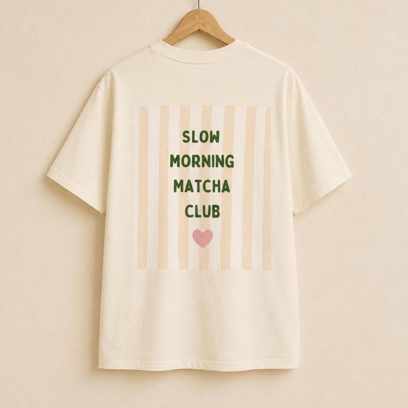 May include: A cream-colored short-sleeve t-shirt featuring a rectangular graphic on the back. The graphic displays vertical beige stripes with the dark green text "SLOW MORNING MATCHA CLUB" and a small pink heart centered below the text.