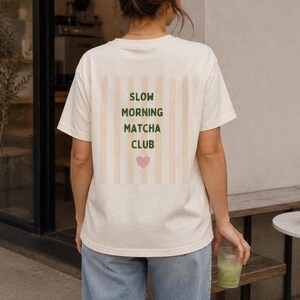 May include: An off-white short-sleeve t-shirt featuring a rectangular graphic on the back. The graphic displays vertical beige stripes with the dark green text SLOW MORNING MATCHA CLUB and a small pink heart icon centered below the lettering.
