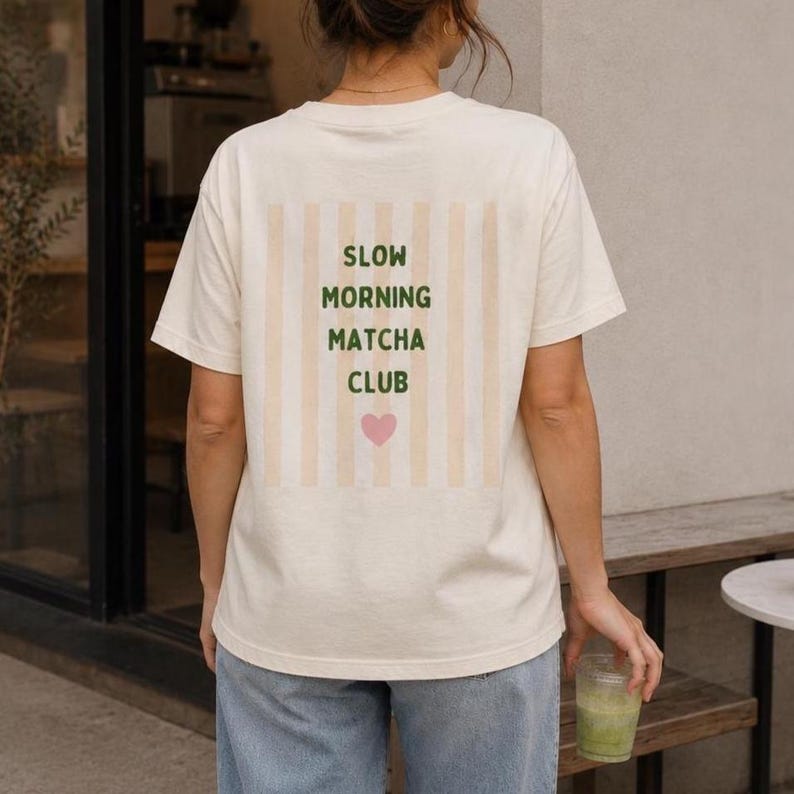 May include: An off-white short-sleeve t-shirt featuring a rectangular graphic on the back. The graphic displays vertical beige stripes with the dark green text SLOW MORNING MATCHA CLUB and a small pink heart icon centered below the lettering.