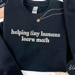 May include: Black sweatshirt with the embroidered text "helping tiny humans learn math." The sweatshirt has a crew neck and long sleeves. A gold necklace with a pendant is draped on the front. The word "Black" is in a circle on the bottom right.