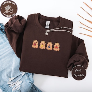May include: Dark chocolate-colored sweatshirt with embroidered cartoon characters wearing hats and holding food items. A silver necklace is draped across the front. The sweatshirt is paired with blue jeans. The text "Dark Chocolate" is in a circle.