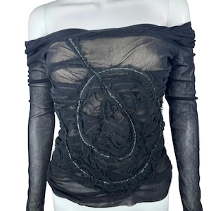 May include: Black off-the-shoulder top with long sleeves. The sheer fabric is gathered and ruched, with a textured design on the front. Silver stitching accents the design, adding a subtle shimmer. The top is displayed on a white mannequin.