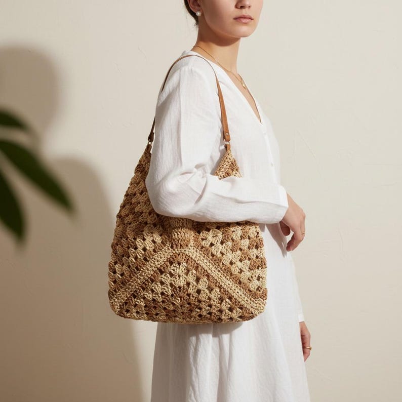 May include: A beige and tan crochet tote bag with brown leather straps. The bag has a geometric pattern and is carried over the shoulder. The woman is wearing a white dress.