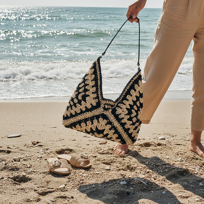 May include: A black and beige crochet tote bag with a black strap is held up on a sandy beach. The bag has a geometric pattern. Beige sandals with a gold buckle are on the sand near the bag.