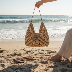 May include: A beige and brown crocheted tote bag with a geometric design is suspended by a hand, featuring brown leather straps. Sandals with buckles rest on the sand. The background shows a beach and the sea.